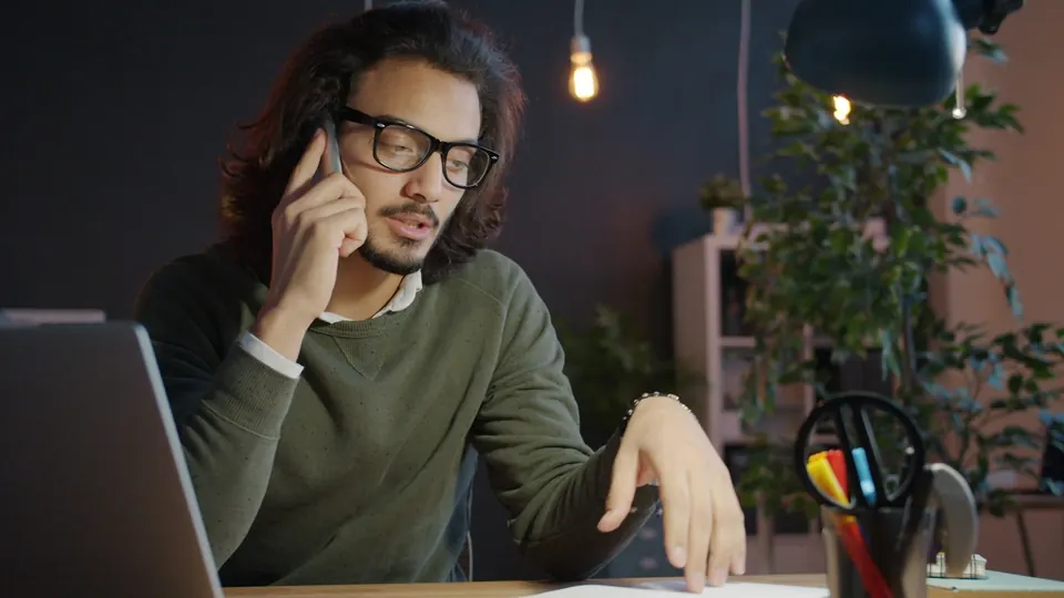 Homem de óculos fala ao celular à noite no escritório, com o notebook aberto